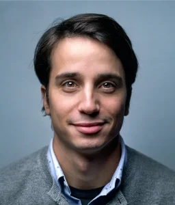Portrait of a young man with dark hair and brown eyes, wearing a gray sweater over a collared shirt, smiling against a light blue background.