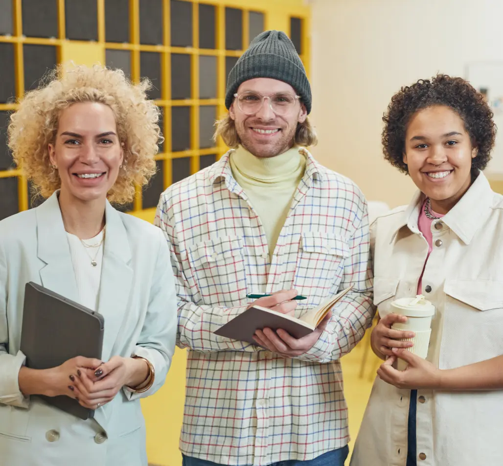 Diverse group of three young professionals smiling together in a modern office setting, with one holding a notebook, another a tablet, and the third a coffee cup, showcasing collaboration and creativity in a vibrant workspace.
